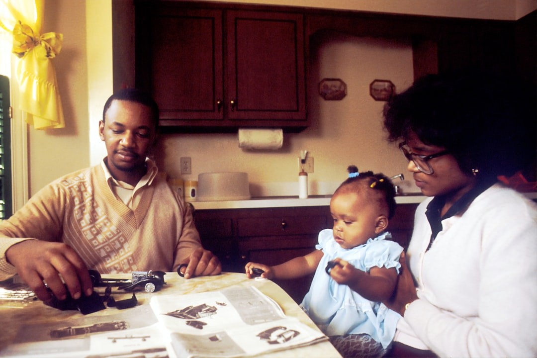 Family Sitting at Kitchen Table. An African American man with his wife and daughter at the kitchen table. He is assembling a Rolls Royce model car indicating the digital facility he maintains. He was diagnosed as having osteogenic sarcoma in his upper left arm. Surgeons performed a new procedure, implanting a metal rod in place of the cancerous bone. Since the muscle and tendon were saved he has the use of this arm.